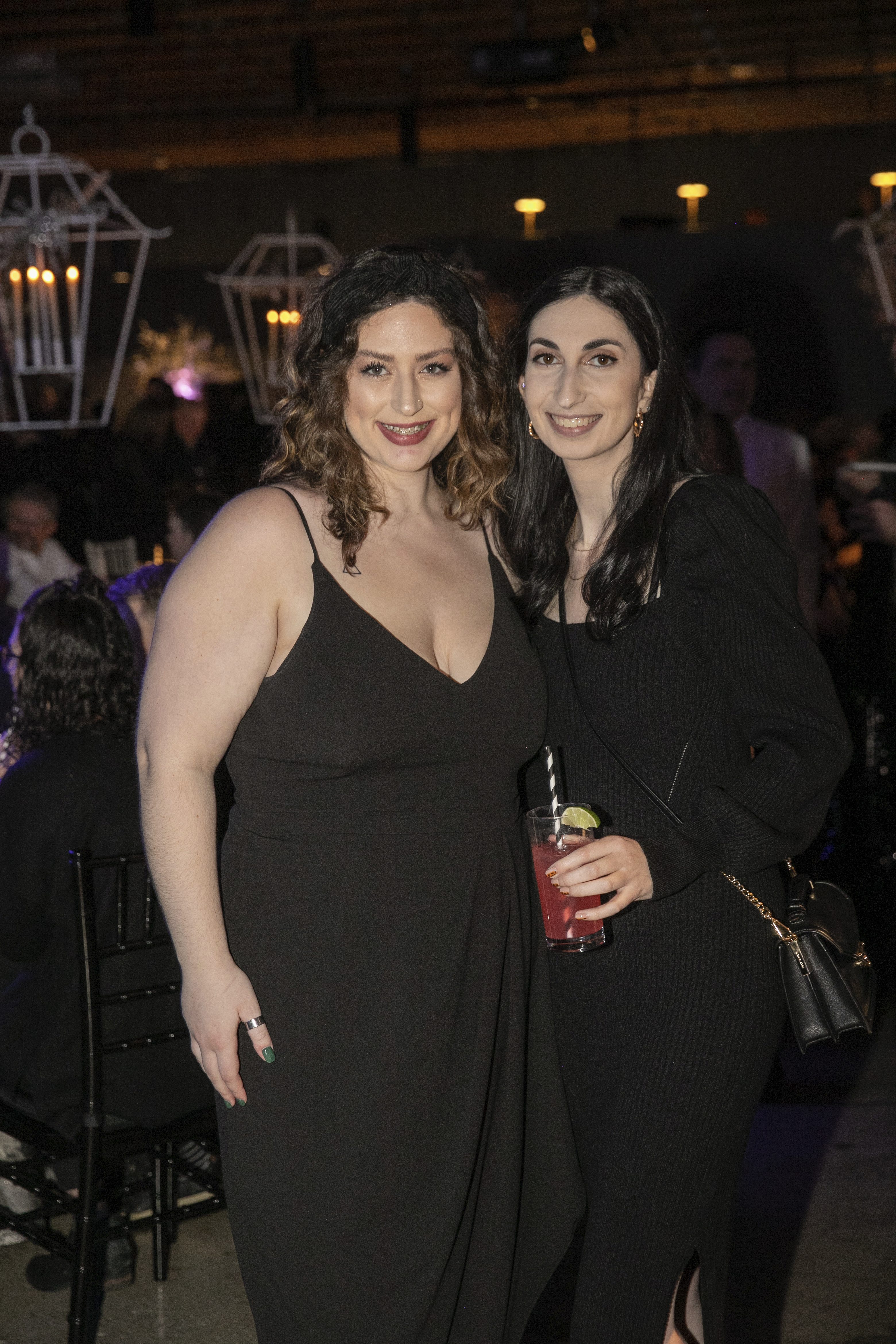 Two women in black fancy clothes, one with drink in hand, smiling at camera